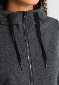 Sudadera con cremallera de color gris oscuro con una textura suave, que presenta una cremallera y cordones en negro contrastante, además de un diseño de cuello alto.