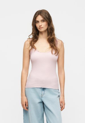 Mladá žena s dlhými vlnitými hnedými vlasmi nosí svetloružový tielkový top a svetlomodré široké džínsy, stojí pred jednoliatym bielym pozadím.