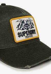 Oliwkowa czapka z kremową naszywką przedstawiającą góry, z logo "SUPERDRY" oraz żółtą bordiurą; zawiera lekko zużytą daszek.