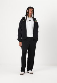 Sweatshirt preto com fecho e logótipo, t-shirt branca com texto bordado, calças de fato de treino pretas e ténis brancos com detalhes cinzas.