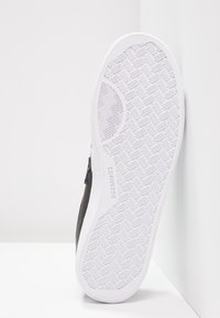 Semelle en caoutchouc blanche d'une chaussure Converse montrant un motif de semelle détaillé, inclinée contre un mur et une surface blanche.