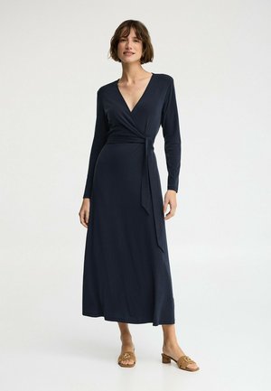 Vrouw draagt een marineblauwe wikkeljurk met lange mouwen en een strik aan de zijkant, gecombineerd met beige slippers, staand tegen een effen lichte achtergrond.