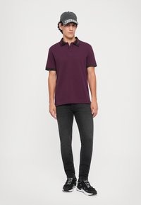 Camisa polo bordô com colarinho e mangas pretas estampadas, combinada com jeans escuros e sapatilhas de desporto pretas. Design simples e moderno.