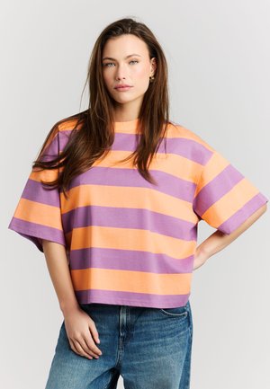 Donna con lunghi capelli castani indossa una camicia oversize a maniche corte con larghe strisce arancioni e viola e jeans blu, in piedi davanti a uno sfondo bianco.