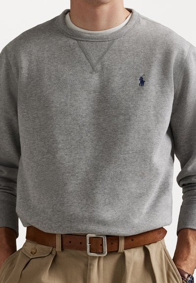 Sweatshirt cinza com gola redonda, punhos canelados e um pequeno logótipo azul no peito. Combinado com calças caqui e um cinto castanho.