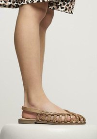 Sandalias trenzadas marrones con punta abierta, que cuentan con un diseño trenzado y una correa en el tobillo. La suela es plana y ligeramente texturizada.