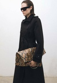 Palton negru tip trench, cu două rânduri de nasturi și curea, asortat cu o poșetă rectangulară, cu un model de nuanțe bej și maro în stil print de șarpe, cu inele din argint.