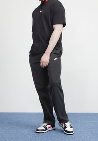 Man standing wearing black Nike Air t-shirt, black Nike sweatpants, and black, white, and red Nike sneakers on blue carpet.