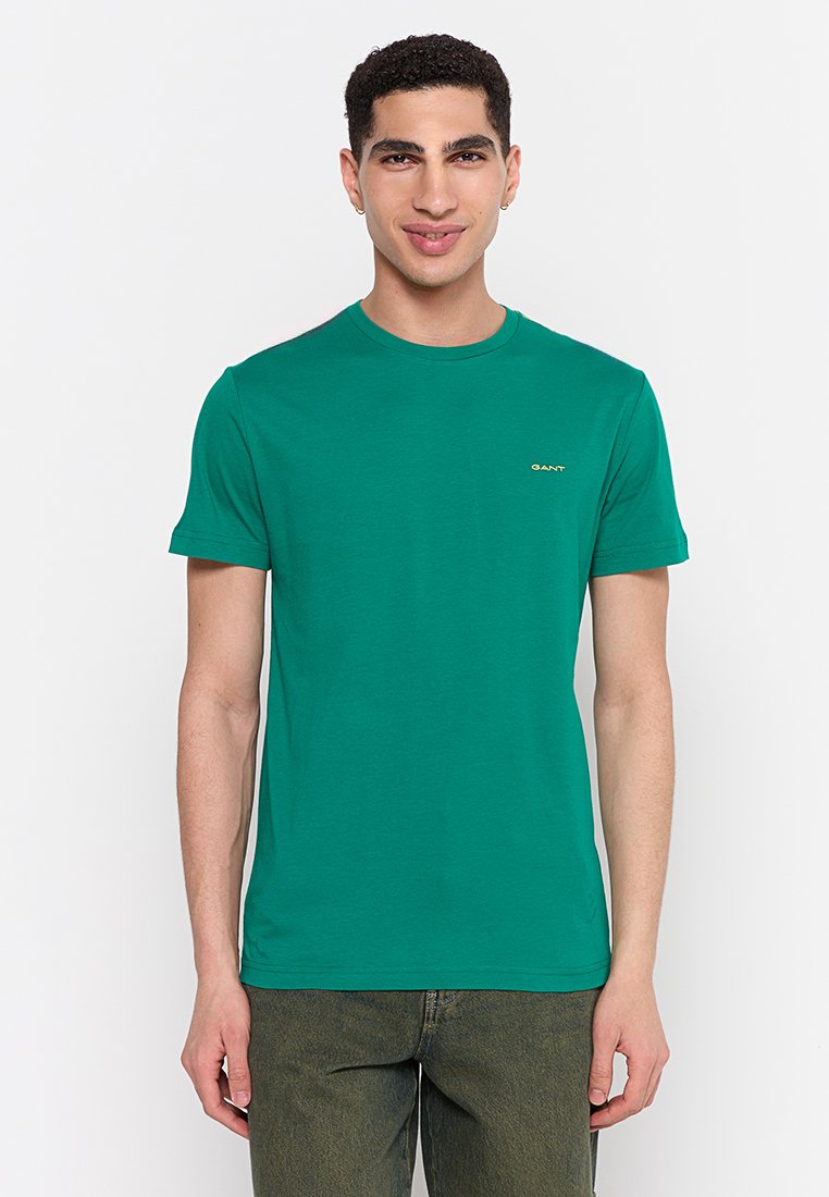 Joven vestido con una camiseta de manga corta verde y jeans oscuros, de pie contra un fondo blanco sencillo, mirando al frente y sonriendo.