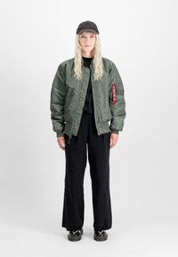 Olijfgroene bomberjack met geribde manchetten, zwart t-shirt, wijde zwarte broek en een donkere pet. Bevat een zijritszak met een label.