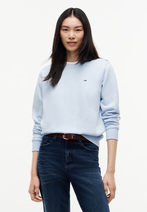 Ung kvinde med lige sort hår iført en lyseblå sweatshirt og mørkeblå jeans, stående mod en ensfarvet lys baggrund.