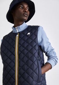 Quilted navy vest with a high collar, zip closure, and two side pockets. Features a multicolored stripe and a small logo on the chest.