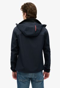 Giacca con cappuccio blu navy scuro realizzata in tessuto liscio, con logo rosso sulla schiena e polsini regolabili. Abbinata a jeans di denim.