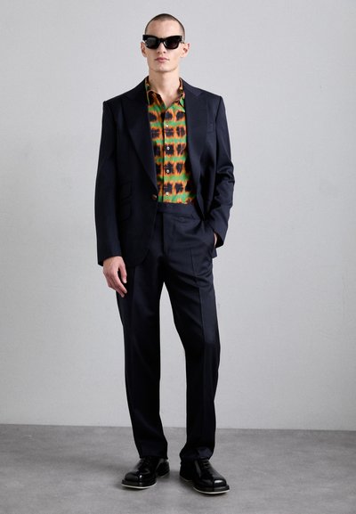 Veste de costume noire ajustée sur une chemise à motifs vifs verts, oranges et noirs. Lunettes de soleil noires, pantalons à jambes larges et chaussures cirées.