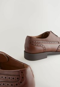Chaussures en cuir marron avec une tige texturée, perforations décoratives et une semelle sombre contrastante. Design à lacets avec un bout arrondi.