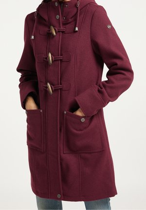 Femme portant un manteau bordeaux à capuche avec des fermoirs à bascule, poches avant, mains dans les poches, et jean bleu.