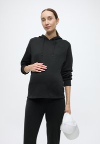 Czarna bluza z kapturem o luźnym kroju w połączeniu z czarnymi spodniami. Model trzyma białą czapkę, ukazując gładką teksturę i prosty design.