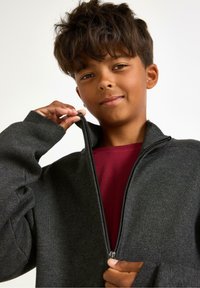 Chlapec s krátkými kudrnatými vlasy, oblečený v vínovém tričku a tmavě šedé mikině se zipem, zapínající zip.