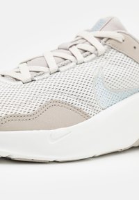 Svijetlosivi tenisice s gornjim dijelom od mrežaste tkanine, bež naglascima i bijelim srednjim dijelom. Ima vezice i suptilni plavi Nike logotip sa strane.