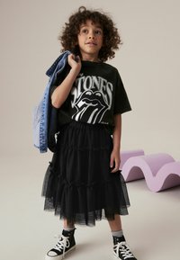 Jupe en tulle noire à volants, associée à un t-shirt graphique noir avec un logo, une veste en denim et des baskets montantes noires.