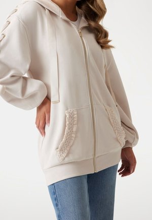 Kobieta w kremowej bluzie z kapturem na suwak z kieszeniami wykończonymi koronką i koronkowymi detalami na rękawach, zestawiona z niebieskimi dżinsami.
