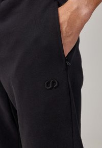 Zwarte sport joggers met een gladde textuur, voorzien van een ritszak en een geborduurd logo aan de zijkant.