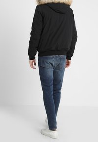 Muž oblečený v tmavě modrých džínách, bílých teniskách a černé bundě s kapucí lemovanou kožešinou, stojící zády k divákovi na jednoduchém pozadí.