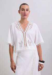 Blouse en crochet blanc avec un décolleté en V profond, des manches courtes et des détails en ouvertures. Présente des motifs élaborés et des bords festonnés.