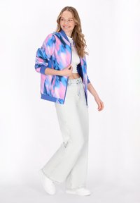 Giubbotto bomber con un motivo astratto blu e rosa, dotato di polsini a coste, chiusura con zip e vestibilità rilassata. Indossato con pantaloni in denim chiaro e sneakers bianche.