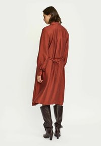 Roodbruine, langemouw jurk van een gestructureerde stof, met een ceintuur in de taille en een vloeiende silhouet. G draagt het met kniehoge bruine laarzen.