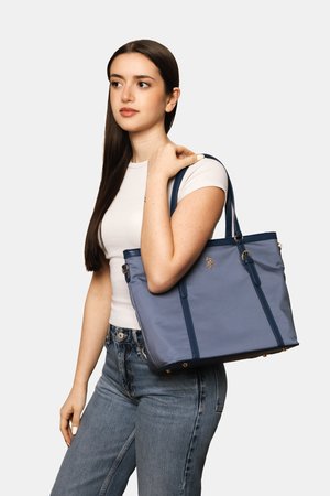 Junge Frau mit langen dunklen Haaren steht und hält eine blaue Tragetasche auf der Schulter, trägt ein weißes T-Shirt und blaue Jeans.