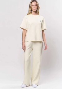 Femme portant un haut ample crème à manches courtes et un pantalon large avec des chaussures blanches, debout devant un fond clair uni.