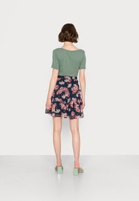 Nainen, jolla on lyhyet ruskeat hiukset, pukeutuneena ribattuun vihreään paitaan, tummansiniseen kukkakuosiseen hameeseen ja vihreisiin lohkopohjaisiin sandaaleihin, seisoo kasvot valkoista seinää kohti.