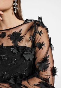 Zwarte doorzichtige blouse met bloemenbroderie, voorzien van een textuur overlay en ingewikkelde bloemenapplicaties langs de halslijn en mouwen.