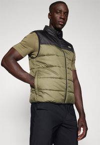 Gilet sans manches avec un panneau supérieur noir et un corps vert olive, fermeture zippée et design léger. Chemise à manches courtes en dessous.