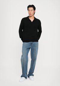 Pulover polo negru cu mânecă lungă și guler plat, purtat cu blugi evazați de culoare bleumarin deschis și adidași albi, stând pe un fundal neutru.