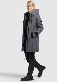 Grå polstret parka med hætte, lynlås lukning, skrålommer og kontrasterende sorte cuffs. Et par sorte knæhøje støvler med teksturerede såler.