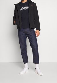 Svart jacka, marinblå t-shirt med grafik, mörka denimjeans, vita sneakers, strukturerad tyg, avslappnad passform, dragkedjestängning på jackan.