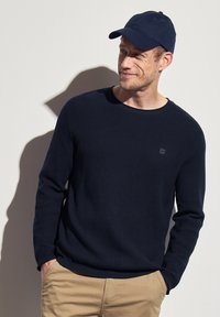 Pull en maille bleu marine avec un col rond, associé à une casquette de baseball bleu marine. Pantalons marron clair. Détail de logo subtil sur le pull.