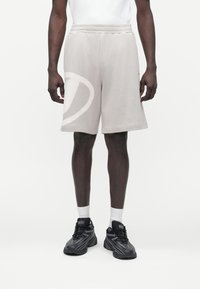 Moški v bež kratkih hlačah do kolen, belih nogavicah in črnih športnih copatih Diesel, stoji pred enobarvnim belim ozadjem.