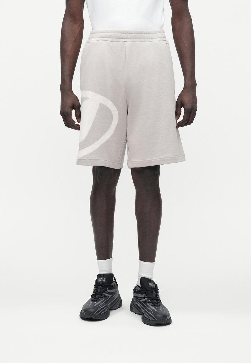 Moški v bež kratkih hlačah do kolen, belih nogavicah in črnih športnih copatih Diesel, stoji pred enobarvnim belim ozadjem.