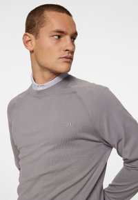 Grå tröja med ribbad halsringning och raglanärmar, prydd med en subtil logotyp på bröstet, tillverkad av ett mjukt material.