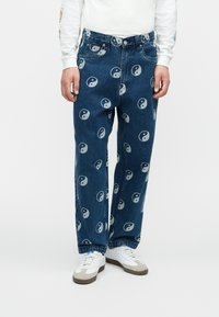 Traperice s ležernim krojem, ukrašene ponovljenim yin-yang uzorkom u svijetloplavoj boji na srednje plavoj pozadini. Osvježene klasičnim dizajnom s pet džepova.