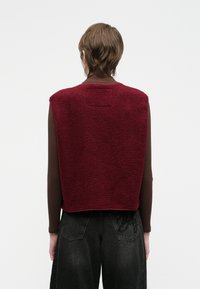 Osoba s kratkom smeškom smešnom frizurom koja nosi bordo flis prsluk preko smešnog dugog rukava i crne izbledele traperice, okrenuta leđima.