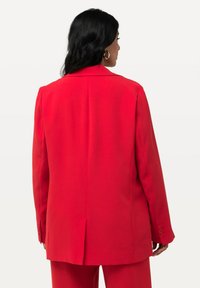 Blazer rosso con un design strutturato, spacco singolo e polsini con quattro bottoni. Tessuto morbido, vestibilità sartoriale e linee pulite.