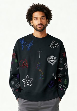 Homme avec barbe et cheveux bouclés portant un sweat-shirt noir décoré de symboles brodés colorés incluant des étoiles, un crâne, un cœur, une croix et des oiseaux.