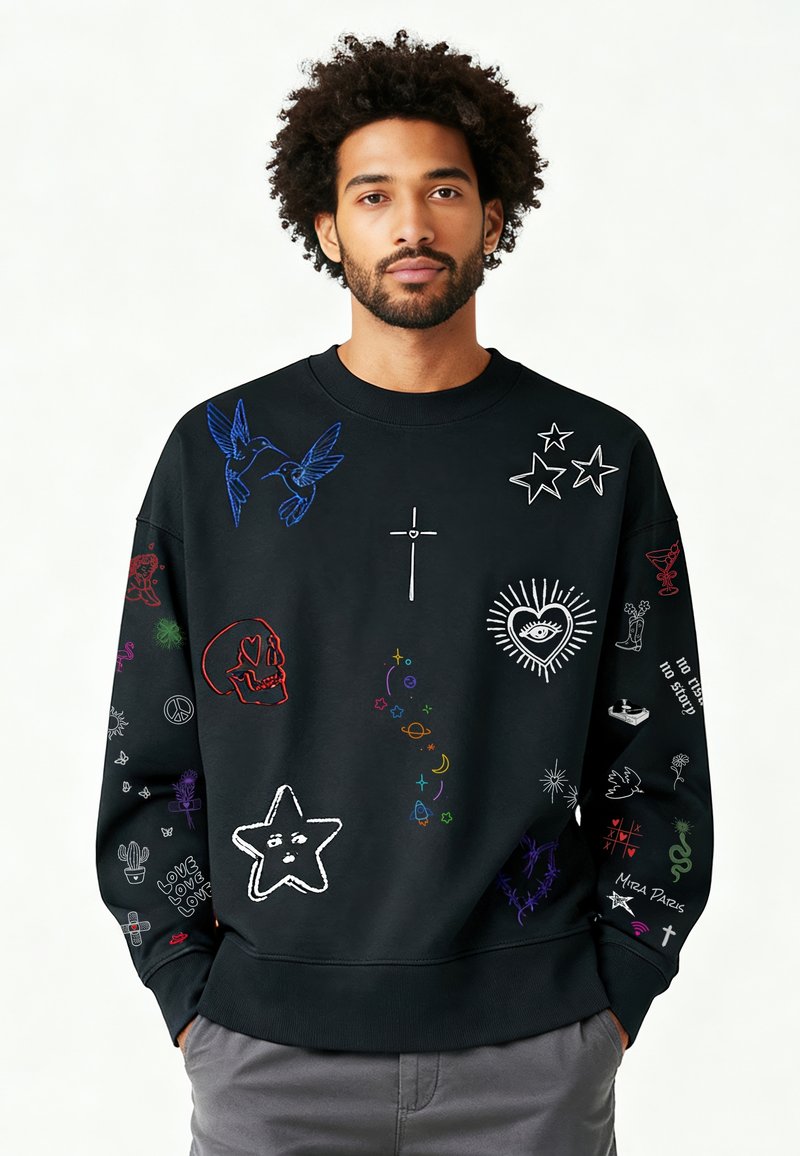 Homme avec barbe et cheveux bouclés portant un sweat-shirt noir décoré de symboles brodés colorés incluant des étoiles, un crâne, un cœur, une croix et des oiseaux.