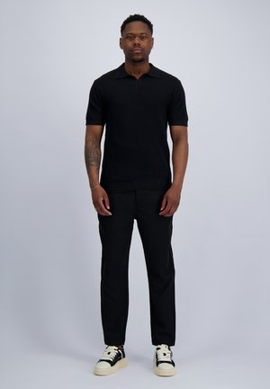 Mann steht und trägt ein schwarzes Kurzarmshirt, schwarze Hose und schwarz-weiße Sneaker vor einem einfarbig hellen Hintergrund.
