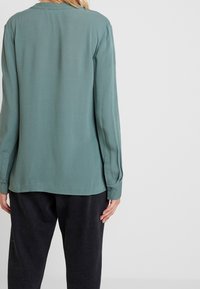 Eine Person, die eine langärmlige türkisfarbene Bluse und lockere schwarze Hose trägt, von hinten vor einem schlichten weißen Hintergrund gezeigt.