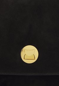Svart mockaväska med ett runt guldmetallspänne med graverad logotyp. Ytan är slät och designen är minimalistisk.
